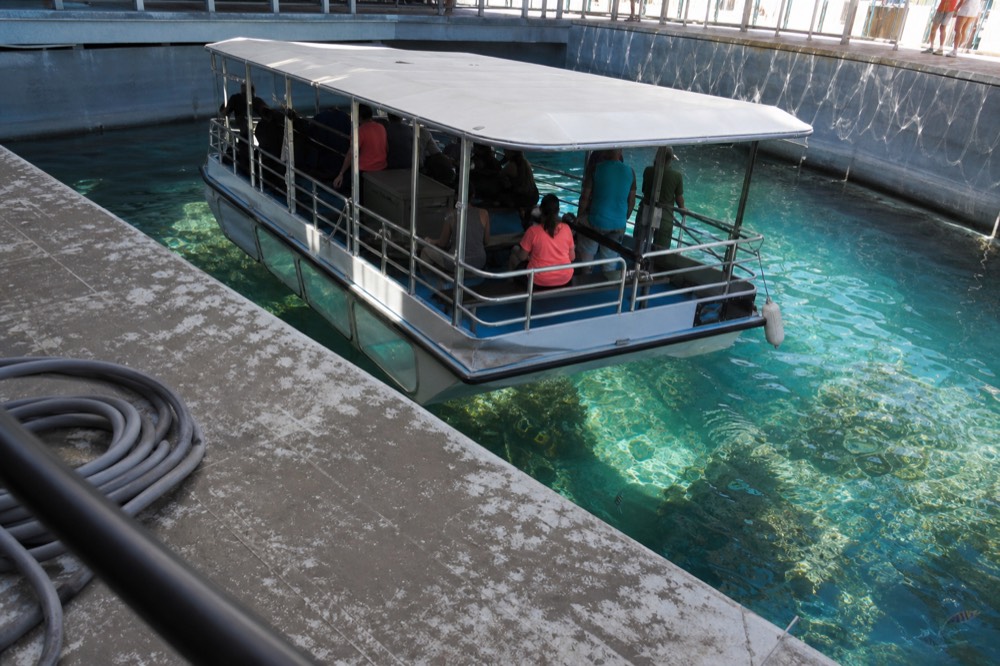 A glass-bottom boat floating over a coral reef tank with fish visible through the hull, no text, no logos, no people