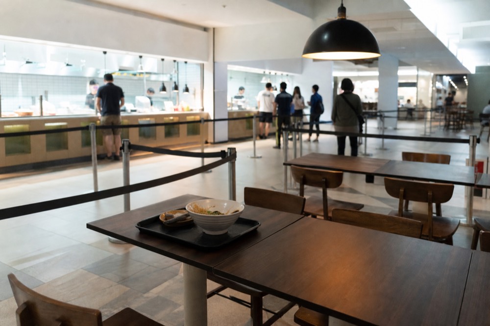 Basement level of a shopping mall with a food court seating area and a queue barrier, no text, no logos, no people.