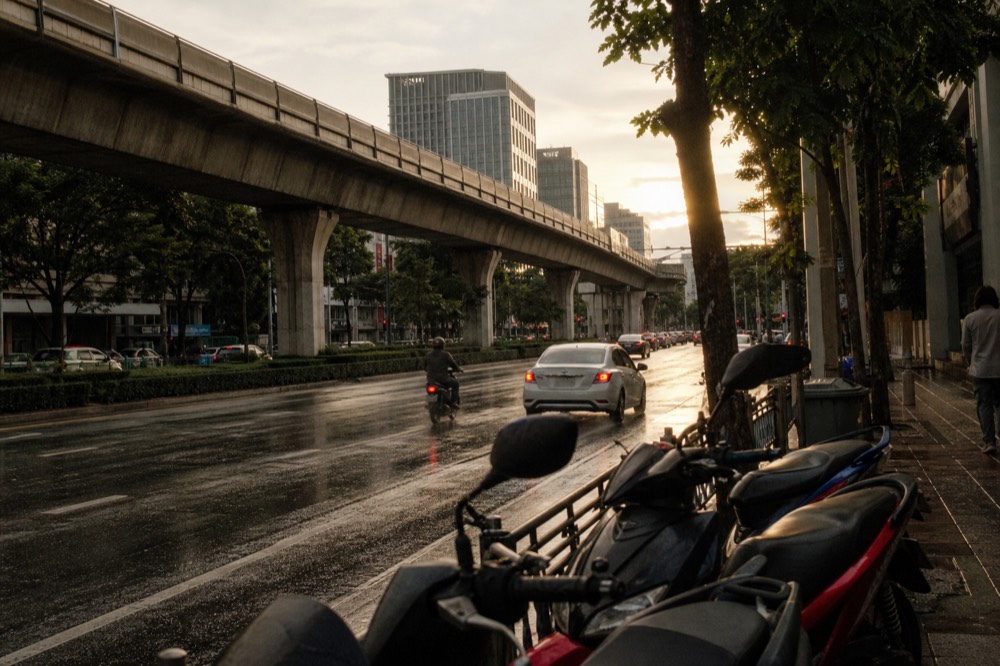 Evening light over a shopping district with a rail line above the road, no text, no logos, no people.